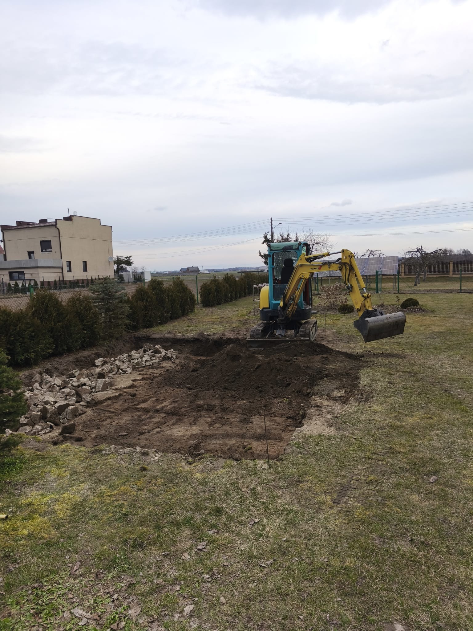 Żółto-niebieska mini koparka w trakcie prac ziemnych na działce, widoczny wykop i sterta kamieni. W tle dom i linia drzew, pochmurne niebo.