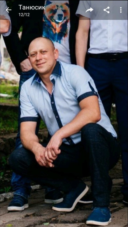 Mężczyzna w jasnej koszuli i ciemnych spodniach kuca na tle innych osób. Uśmiechnięty, z widocznym pierścionkiem na palcu. Nieostre tło z zielenią.