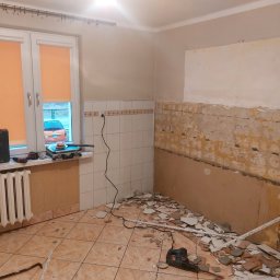 BRK Rafał Kuźdub - Demontaż płytek w kuchni w bloku, widoczne narzędzia, gruz i przygotowane ściany. Okno z pomarańczowymi roletami i skrzynka narzędziowa w tle.