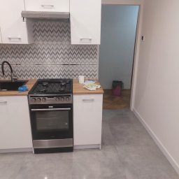 BRK Rafał Kuźdub - Nowoczesna kuchnia w bloku po remoncie: białe szafki, kuchenka Amica, szare płytki chevron na ścianie i podłodze. Widok na otwarte przejście do innego pomieszczenia.