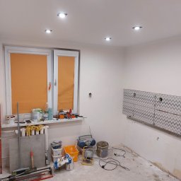 BRK Rafał Kuźdub - Remont kuchni w bloku: częściowo położone płytki z szarym wzorem na ścianie, narzędzia i materiały budowlane w nieukończonym pomieszczeniu. Widoczne światła sufitowe.