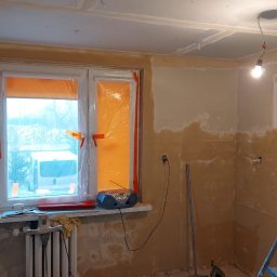 BRK Rafał Kuźdub - Kuchnia w bloku w trakcie remontu: ściany przygotowane pod gładzie, okno zabezpieczone folią, widoczne narzędzia i drabina. Stan surowy przed finalnym wykończeniem.