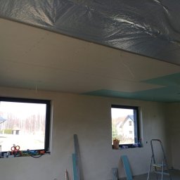 BRK Rafał Kuźdub - Wnętrze w trakcie wykończenia: zamontowana płyta GK na suficie, okna z widokiem na zewnątrz, drabina i narzędzia budowlane. Widoczna folia izolacyjna pod płytami.