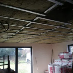 Montaż izolacji z wełny mineralnej na ruszcie sufitowym w domu jednorodzinnym. Widoczne pakiety wełny Rockwool i okno z widokiem na ogród.