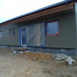 BRK Rafał Kuźdub - Niski dom z szarą elewacją w trakcie prac. Okna i drzwi zabezpieczone folią. Widoczne rusztowania i materiały budowlane. Tło: pole i las.