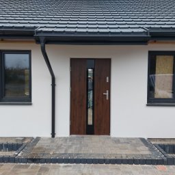 BRK Rafał Kuźdub - Elewacja domu jednorodzinnego z ciemną stolarką okienną i drzwiową, widoczne orynnowanie i wykończone wejście z kostki brukowej. Jedno okno zabite płytą.