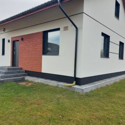 BRK Rafał Kuźdub - Nowoczesny dom jednorodzinny z białą elewacją, drewnianym panelem przy wejściu i czarnymi detalami, takimi jak rynna i podmurówka. Widoczne schody wejściowe i zadbany trawnik.