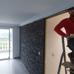 BRK Rafał Kuźdub - Wykończone wnętrze domu: ściana z kamienia dekoracyjnego, szare panele podłogowe i biały sufit z listwami. W tle widoczne okno balkonowe i fragment drabiny.