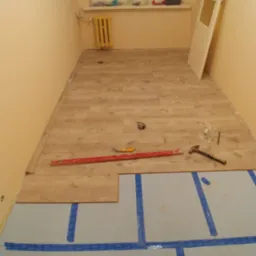 Fragment układania paneli podłogowych w pokoju. Częściowo ułożone panele, narzędzia pracy, podkład z niebieską taśmą. Widok z góry.