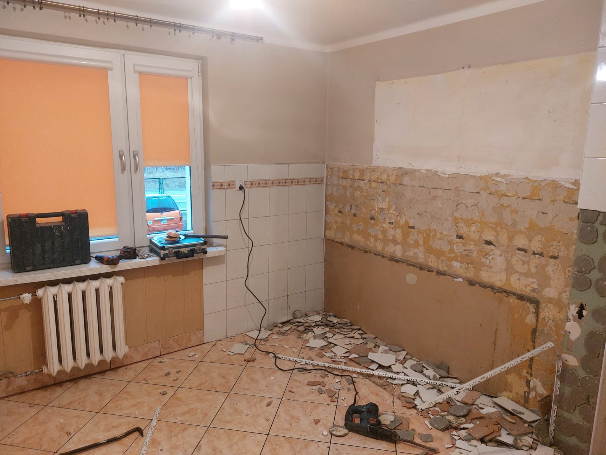 Kuchnia w bloku w trakcie remontu. Usunięte płytki ze ściany, widoczne narzędzia i gruz na podłodze. Okno z pomarańczowymi roletami i grzejnik.