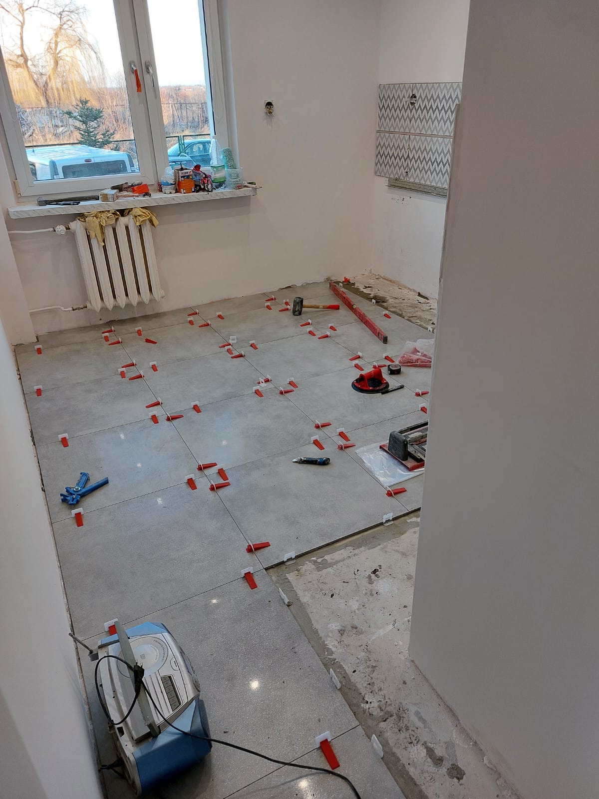 Kuchnia w bloku: etap układania szarych płytek podłogowych z użyciem czerwonych klinów poziomujących. Widoczne narzędzia glazurnicze i fragment ściany z ułożonymi płytkami.