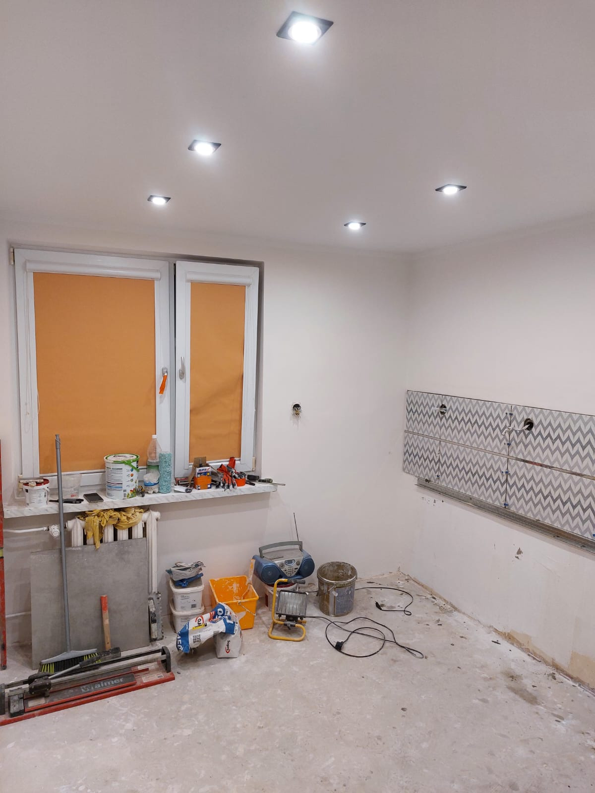 Remont kuchni w bloku: częściowo położone płytki z szarym wzorem na ścianie, narzędzia i materiały budowlane w nieukończonym pomieszczeniu. Widoczne światła sufitowe.