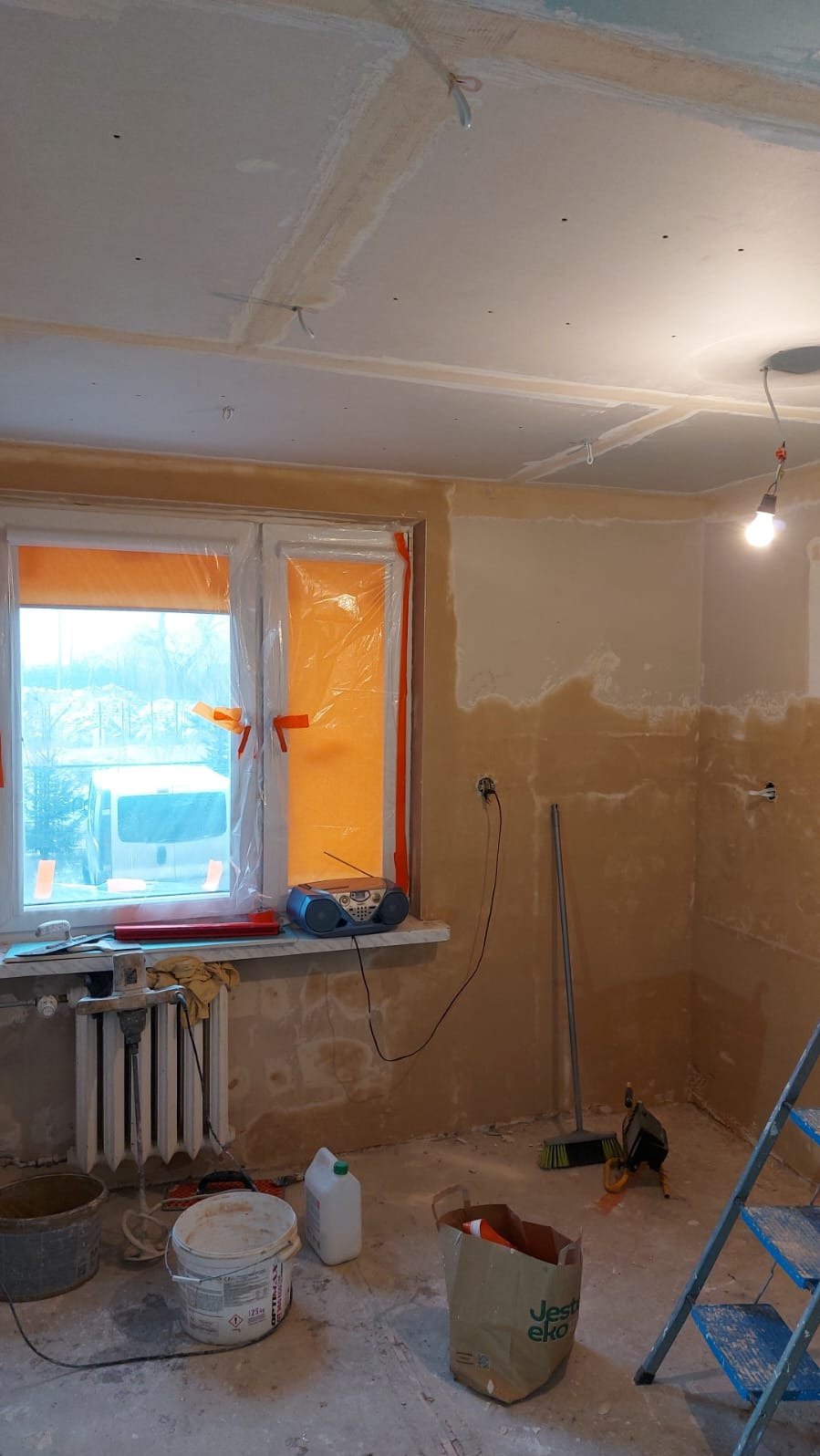 Kuchnia w bloku w trakcie remontu: ściany przygotowane pod gładzie, okno zabezpieczone folią, widoczne narzędzia i drabina. Stan surowy przed finalnym wykończeniem.