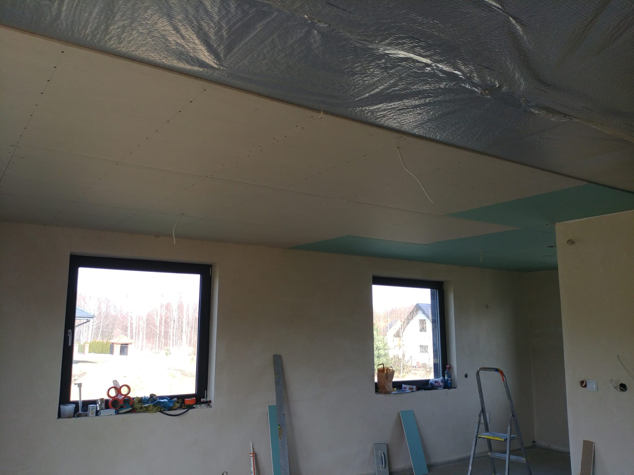 Wnętrze w trakcie wykończenia: zamontowana płyta GK na suficie, okna z widokiem na zewnątrz, drabina i narzędzia budowlane. Widoczna folia izolacyjna pod płytami.