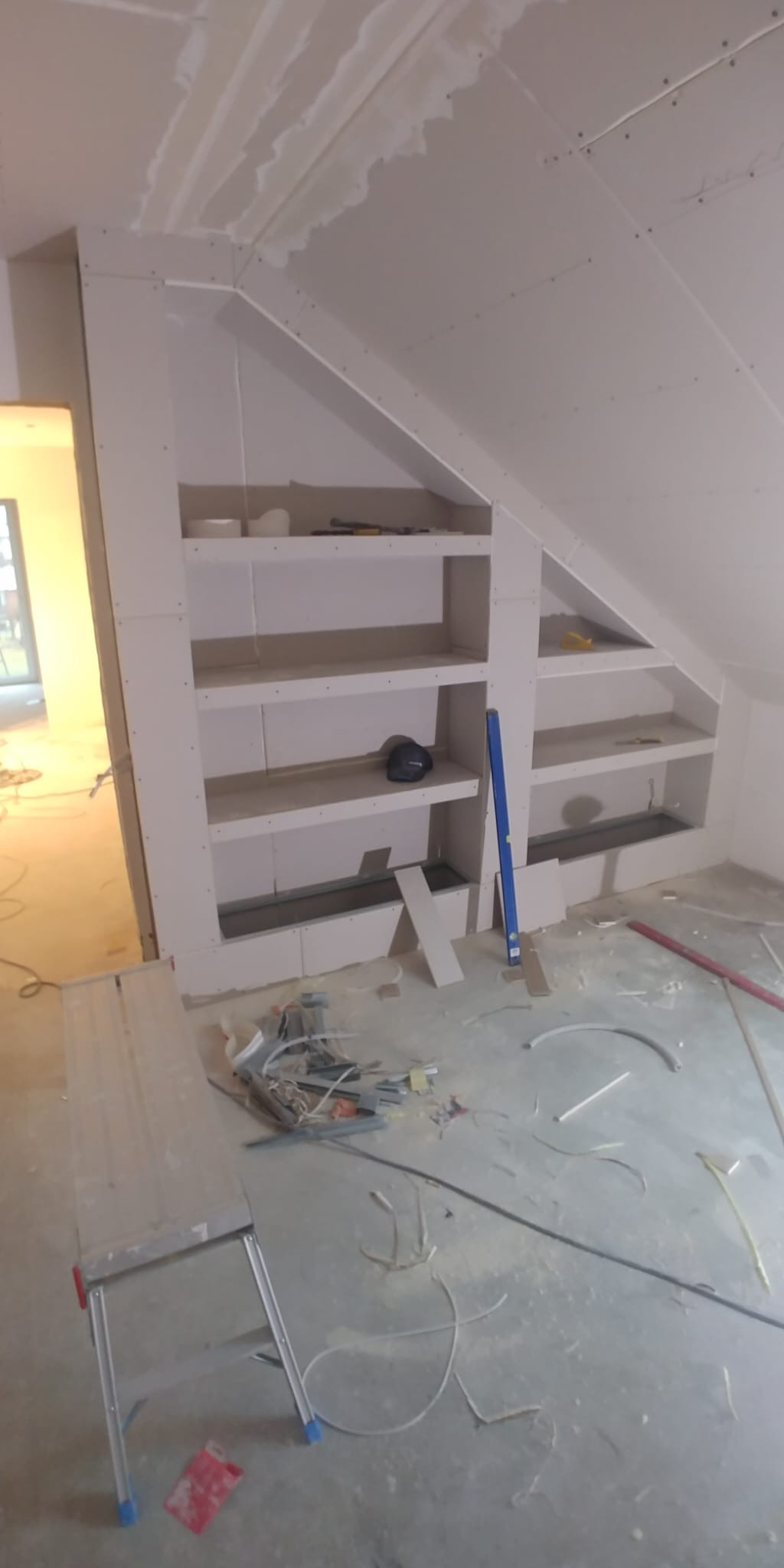 Zabudowa poddasza płytą GK z wnękami na półki. Widoczne narzędzia i resztki materiałów budowlanych. Prace wykończeniowe w domu jednorodzinnym.