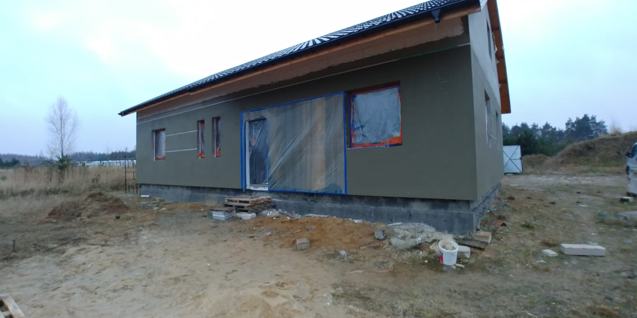 Niski dom z szarą elewacją w trakcie prac. Okna i drzwi zabezpieczone folią. Widoczne rusztowania i materiały budowlane. Tło: pole i las.