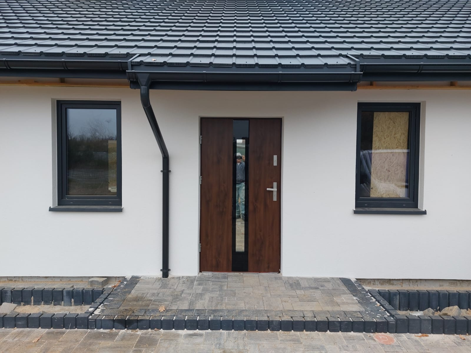 Elewacja domu jednorodzinnego z ciemną stolarką okienną i drzwiową, widoczne orynnowanie i wykończone wejście z kostki brukowej. Jedno okno zabite płytą.