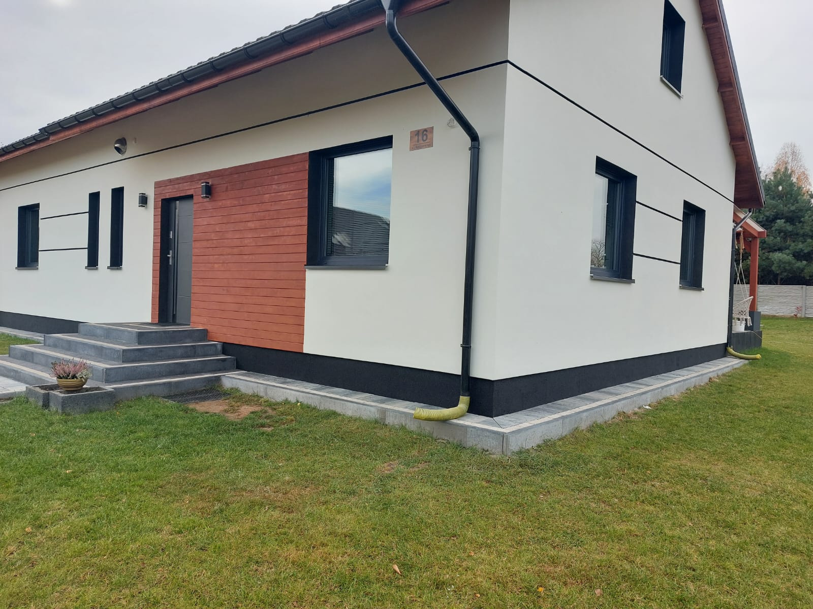 Nowoczesny dom jednorodzinny z białą elewacją, drewnianym panelem przy wejściu i czarnymi detalami, takimi jak rynna i podmurówka. Widoczne schody wejściowe i zadbany trawnik.