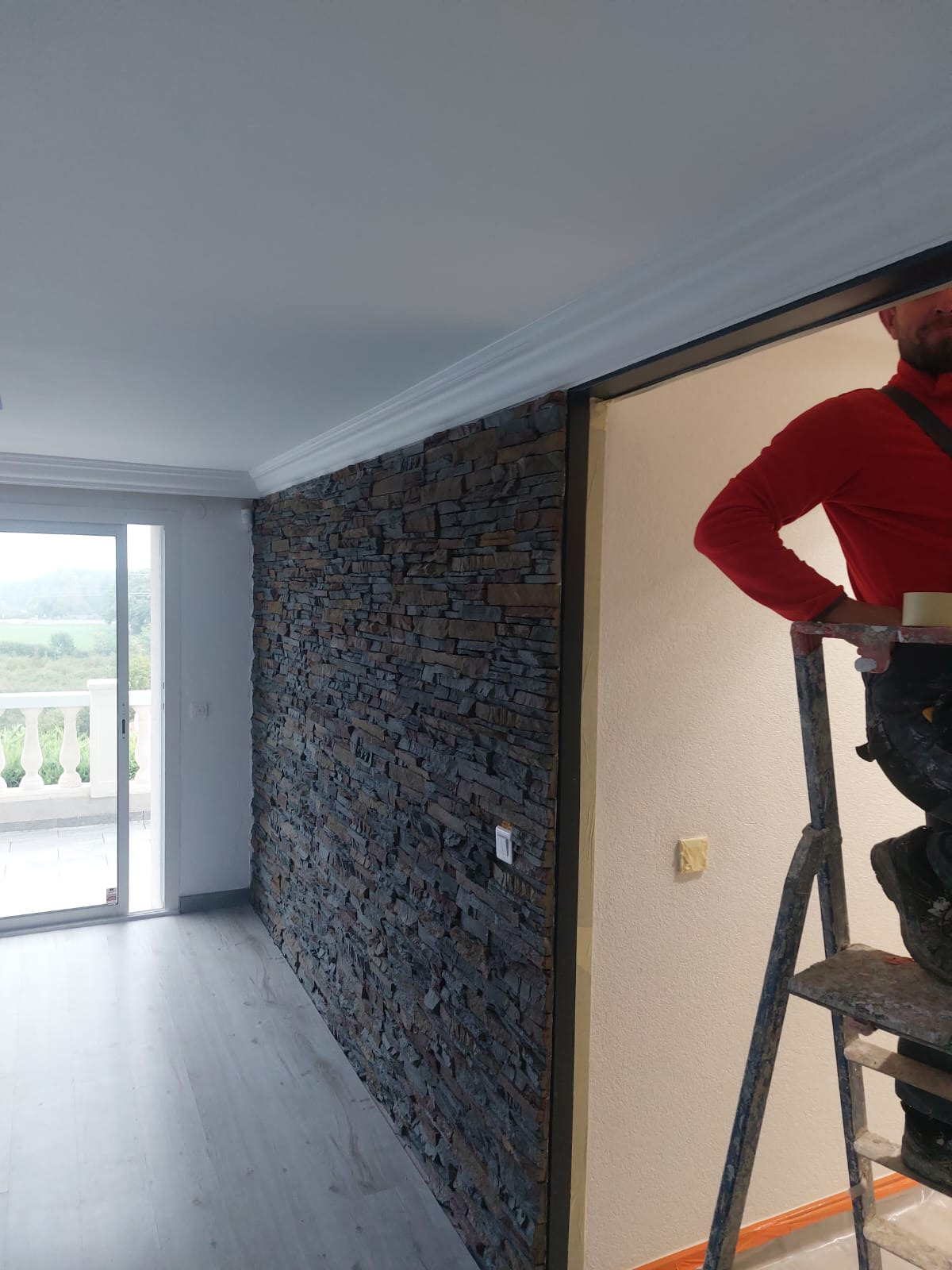 Wykończone wnętrze domu: ściana z kamienia dekoracyjnego, szare panele podłogowe i biały sufit z listwami. W tle widoczne okno balkonowe i fragment drabiny.