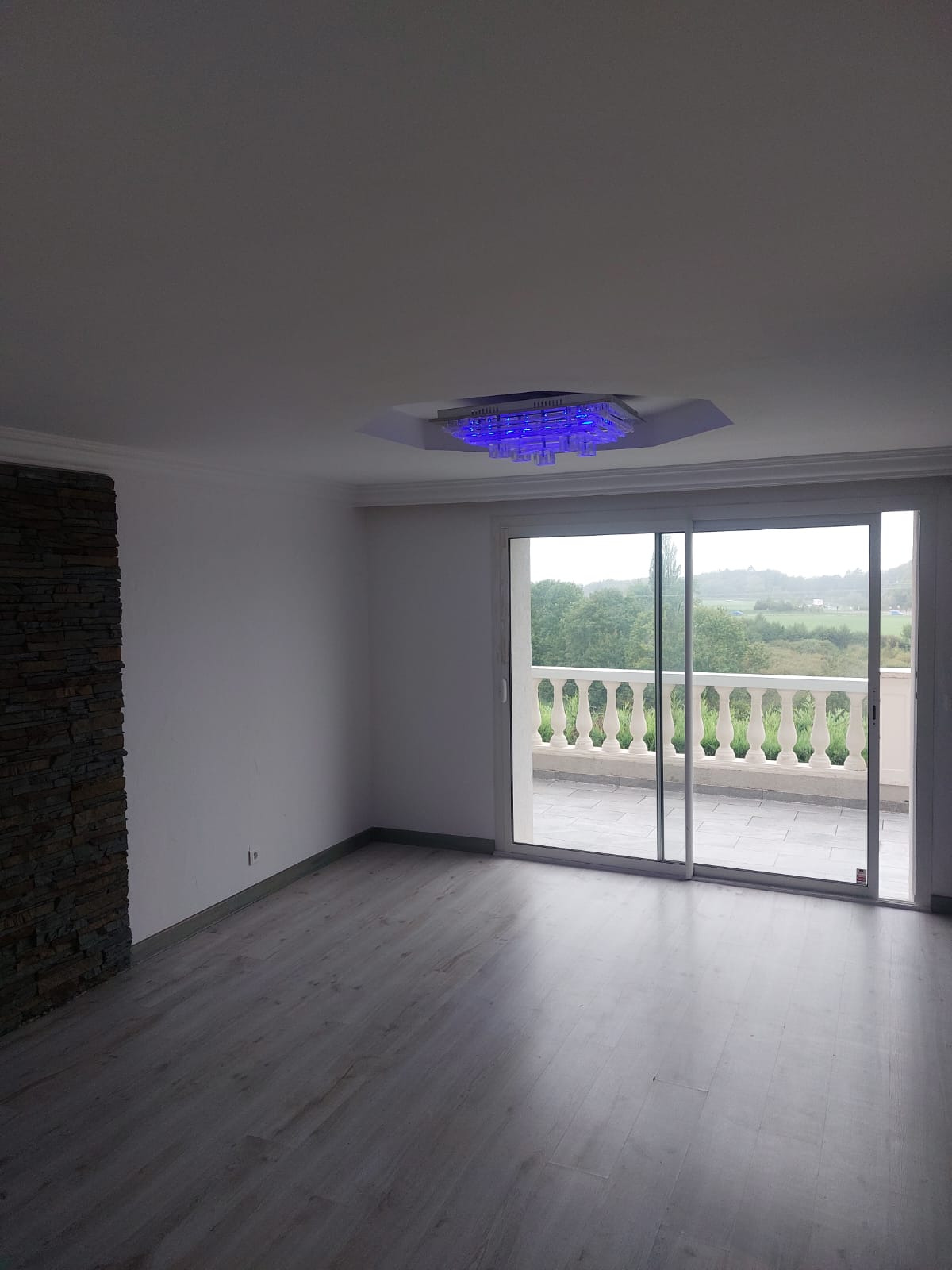 Wykończone wnętrze domu z panelami, ścianą z kamienia dekoracyjnego i balkonem. Widoczny nowoczesny żyrandol z niebieskim światłem. Przesuwne drzwi balkonowe.