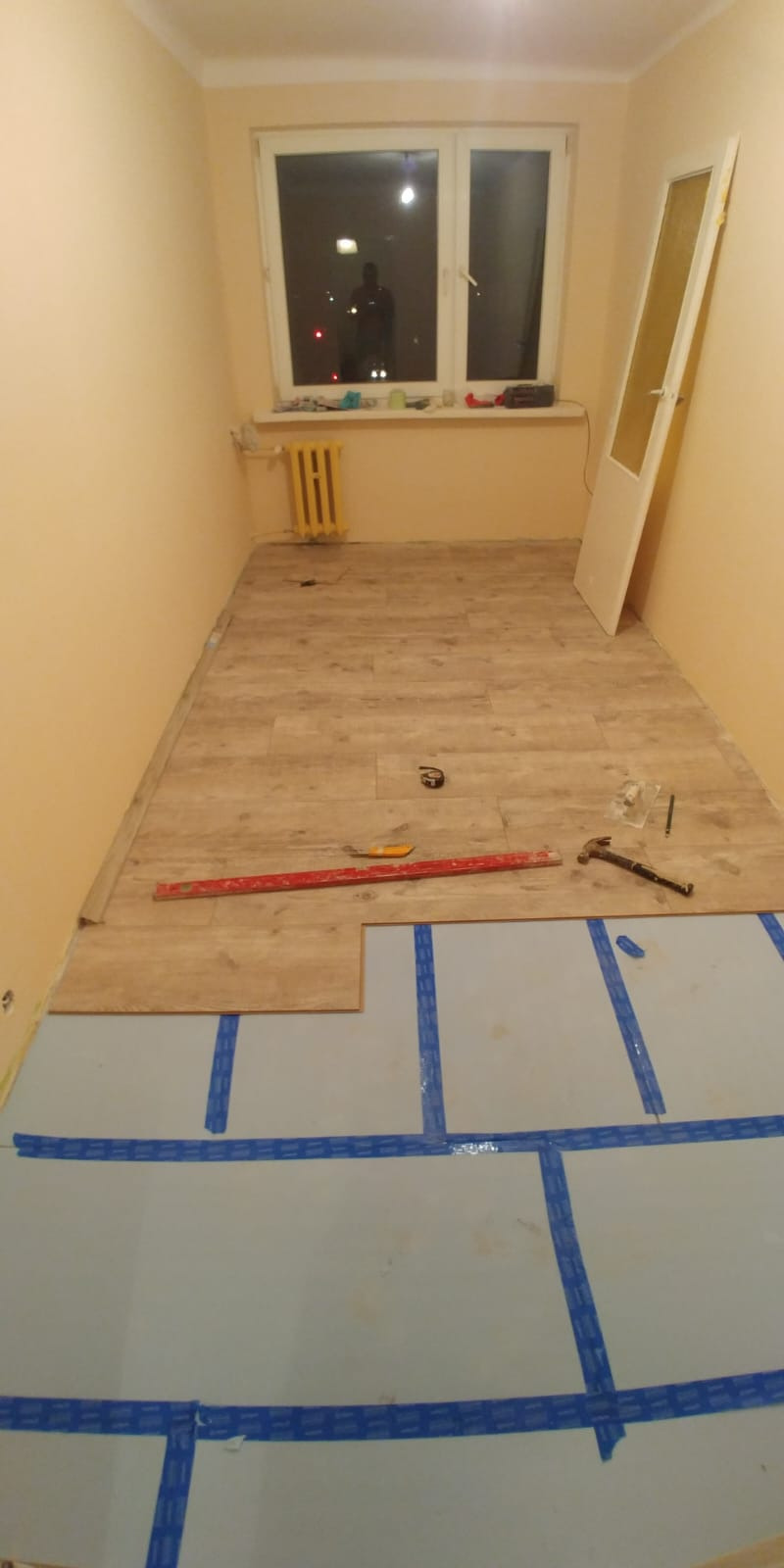 Fragment układania paneli podłogowych w pokoju. Częściowo ułożone panele, narzędzia pracy, podkład z niebieską taśmą. Widok z góry.