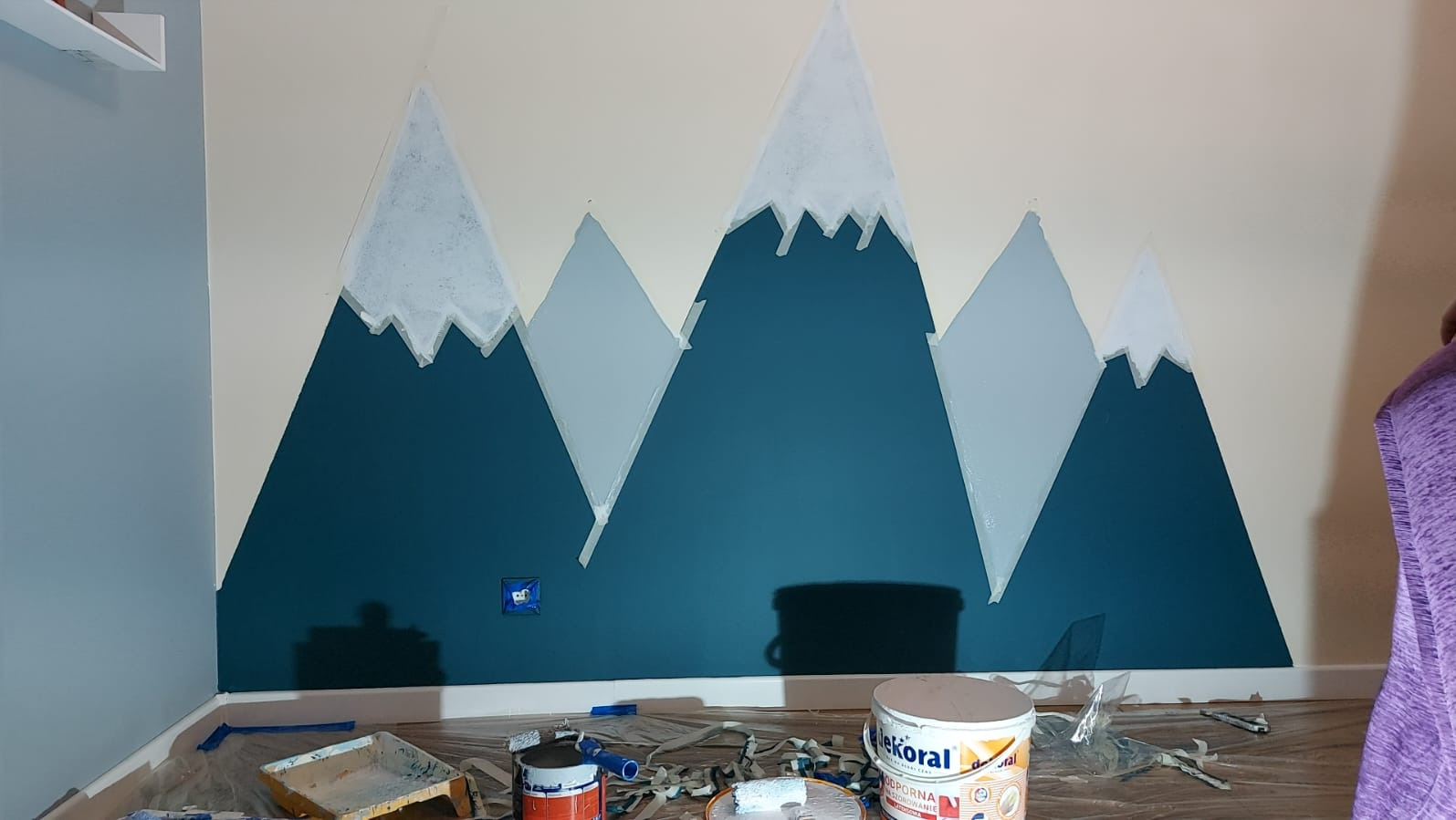 Ściana z motywem gór w odcieniach niebieskiego i szarości, malowana na życzenie klientki. Widoczne puszki z farbą i narzędzia malarskie.
