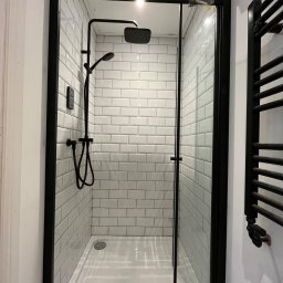 Job On - Kabina prysznicowa z czarną armaturą i białymi płytkami typu metro. Widoczny grzejnik drabinkowy w kolorze czarnym. Nowoczesny design z dbałością o detale.