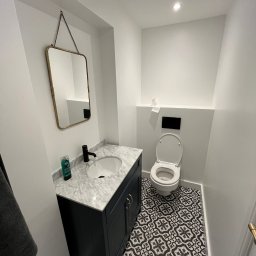 Job On - Kompaktowa łazienka z umywalką na szafce w kolorze granatowym, marmurowym blatem, lustrem w złotej ramie i wzorzystą, czarno-białą podłogą.