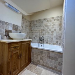 Job On - Jasna łazienka z drewnianą szafką pod umywalką, mozaiką na ścianach i podłodze, białą wanną i oknem dachowym. Styl klasyczny, ciepłe oświetlenie.