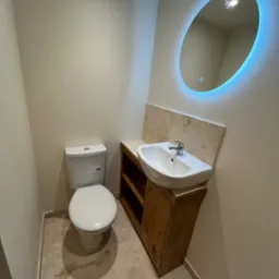 Nowoczesna łazienka z owalnym lustrem LED, jasnymi płytkami i drewnianą szafką pod umywalką. Toaleta kompaktowa w rogu. Aranżacja w stonowanych barwach.