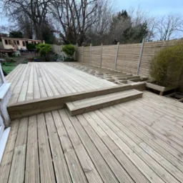 Nowy drewniany taras z niskimi schodami, częściowo w trakcie budowy, z widoczną konstrukcją nośną i drewnianym ogrodzeniem w tle, na zewnątrz budynku.