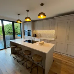 Elegancka, jasna kuchnia z wyspą i drewnianymi stołkami barowymi. Widoczne okno z widokiem na ogród i trzy złote lampy wiszące nad blatem. Nowoczesny design.