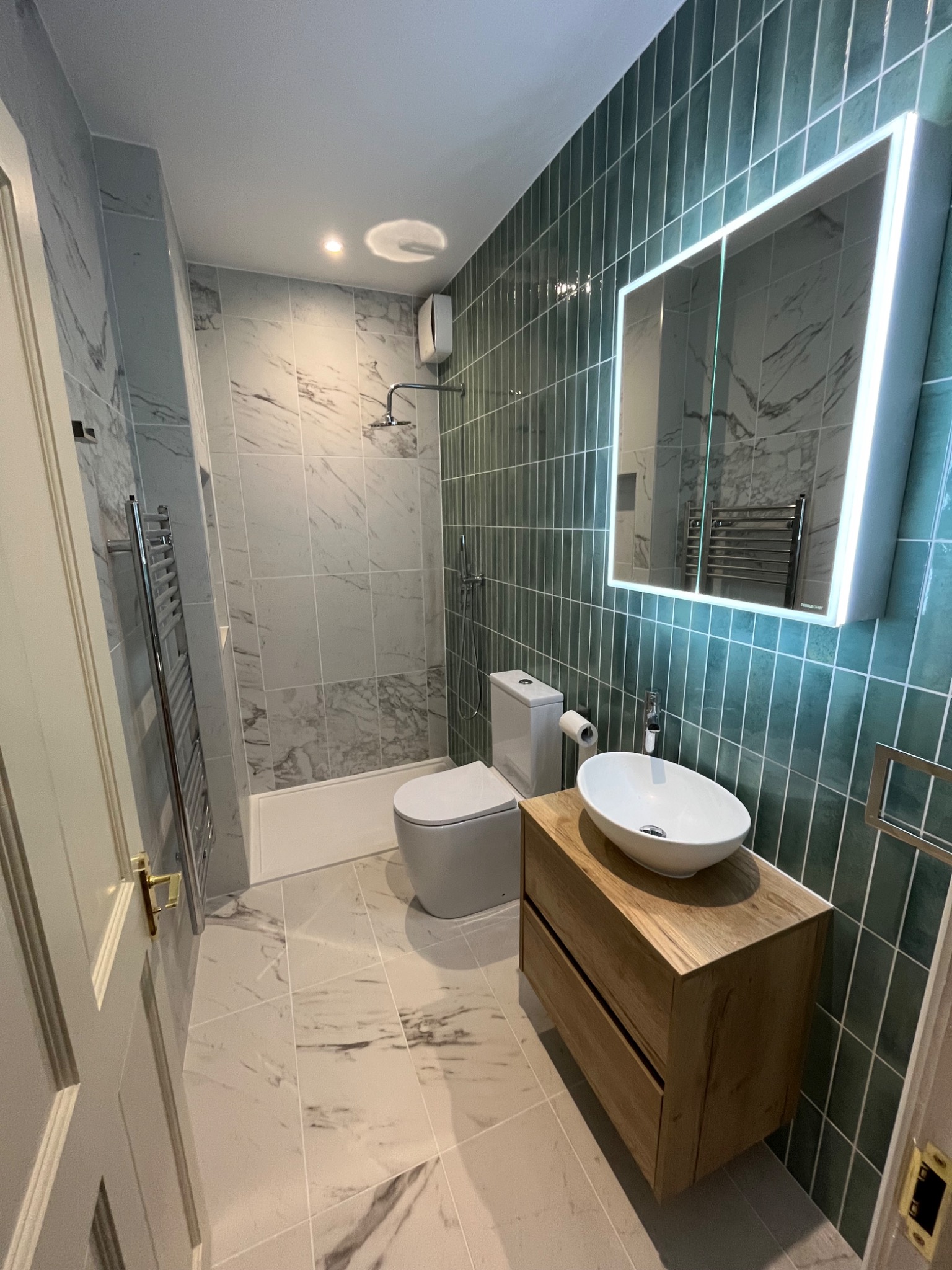 Nowoczesna łazienka po remoncie: zielone płytki typu metro, marmurowe płytki, podświetlane lustro, drewniana szafka pod umywalką i grzejnik drabinkowy.