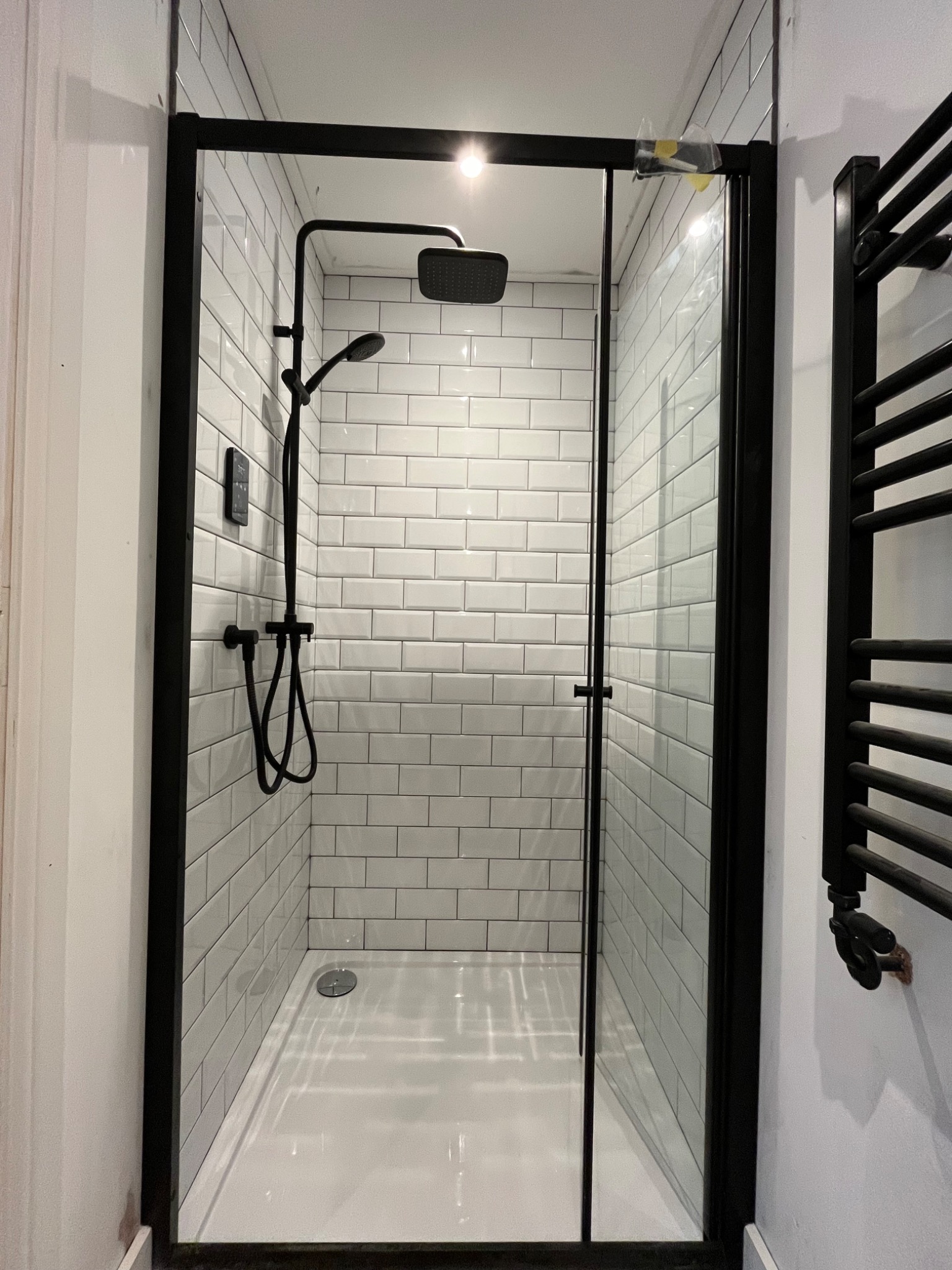 Kabina prysznicowa z czarną armaturą i białymi płytkami typu metro. Widoczny grzejnik drabinkowy w kolorze czarnym. Nowoczesny design z dbałością o detale.