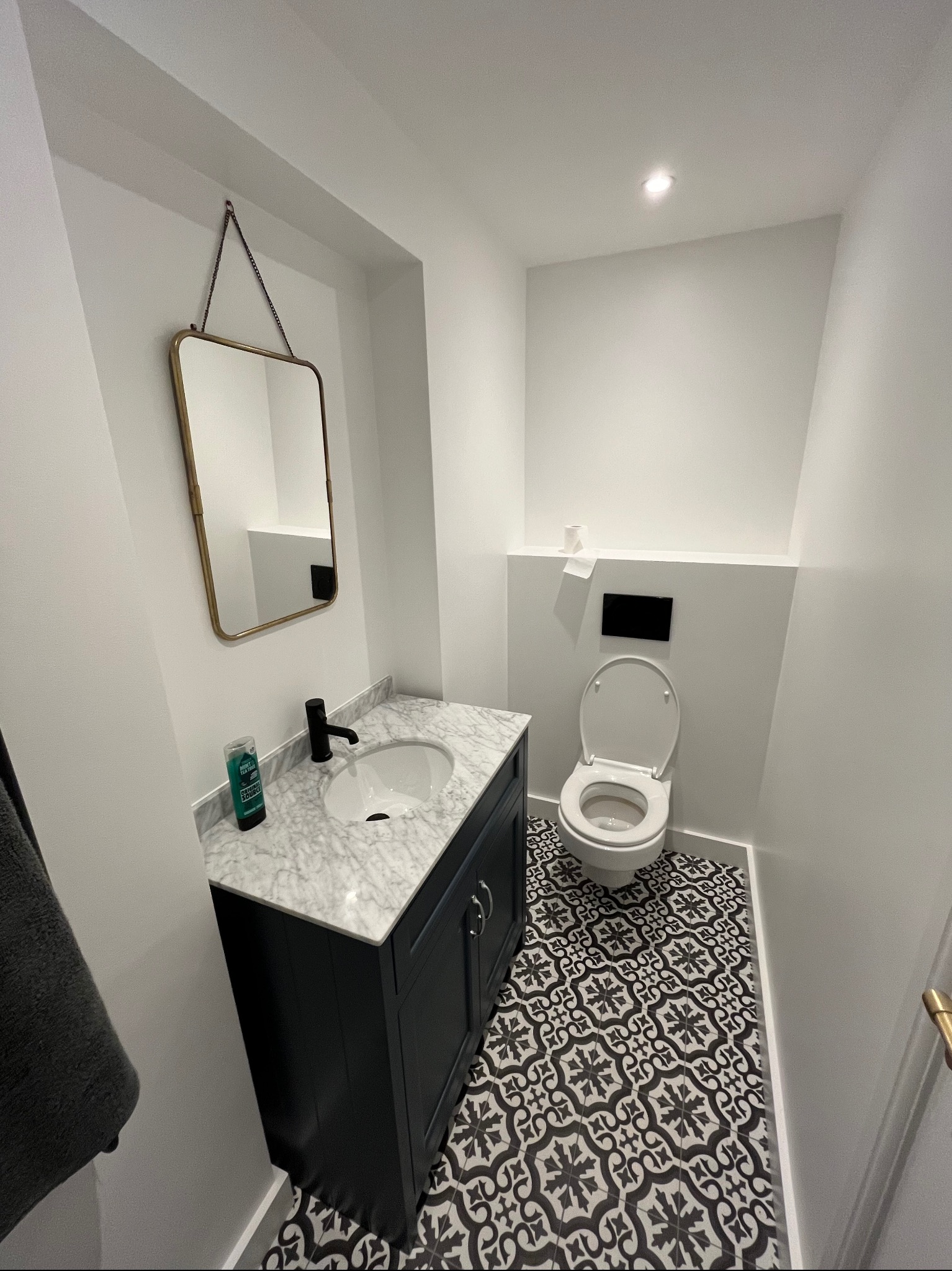 Kompaktowa łazienka z umywalką na szafce w kolorze granatowym, marmurowym blatem, lustrem w złotej ramie i wzorzystą, czarno-białą podłogą.