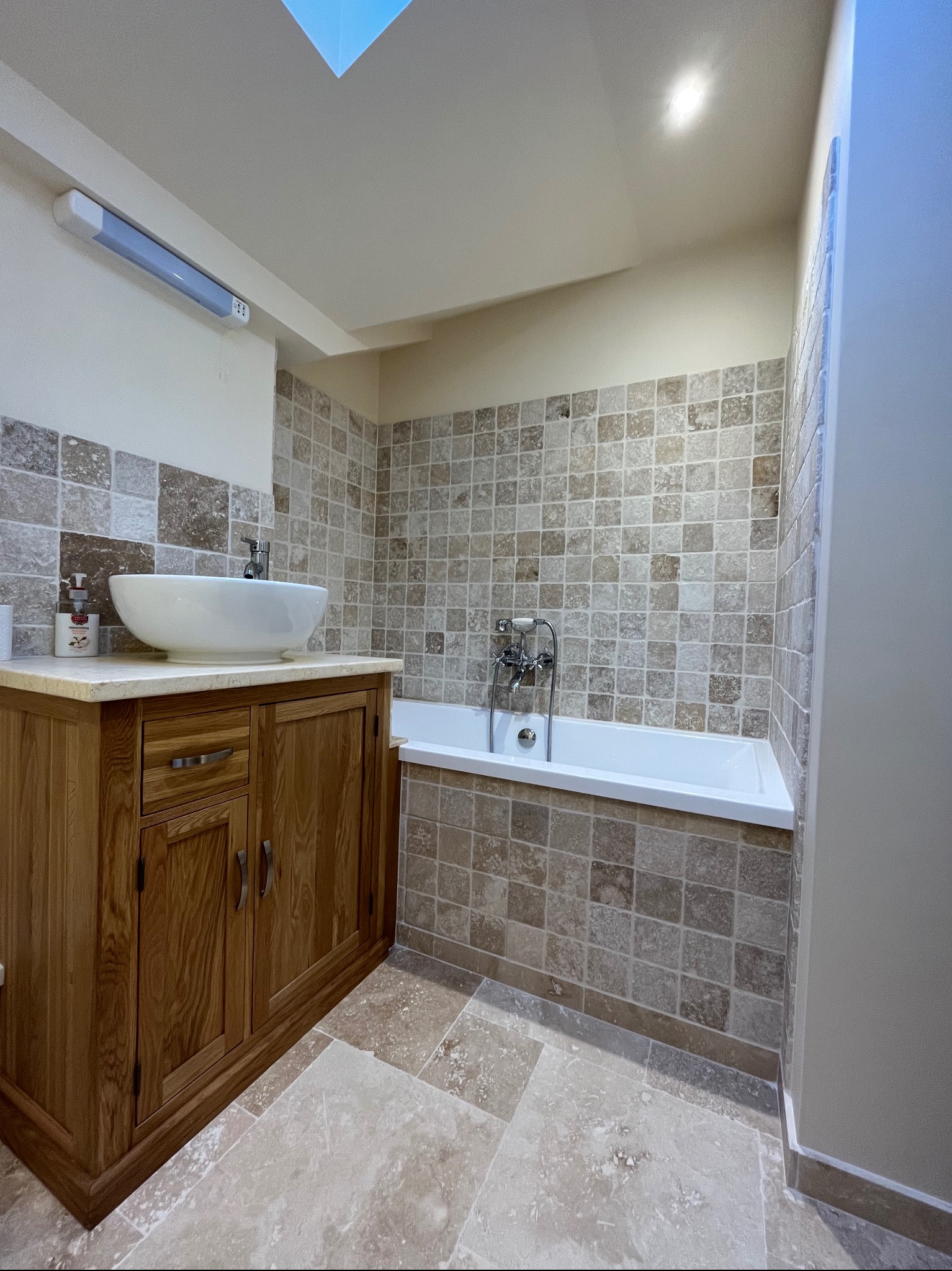 Jasna łazienka z drewnianą szafką pod umywalką, mozaiką na ścianach i podłodze, białą wanną i oknem dachowym. Styl klasyczny, ciepłe oświetlenie.