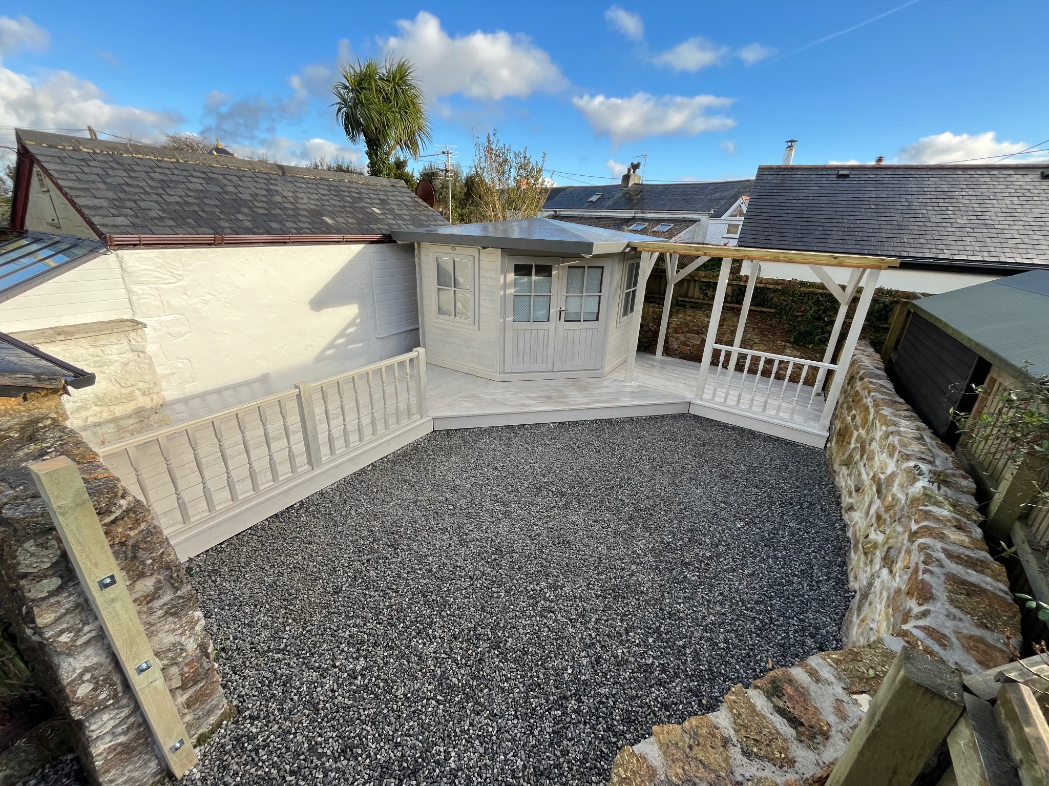 Nowoczesna altana ogrodowa w jasnych barwach, otoczona dekoracyjnym żwirem i kamiennym murkiem. Błękitne niebo w tle.