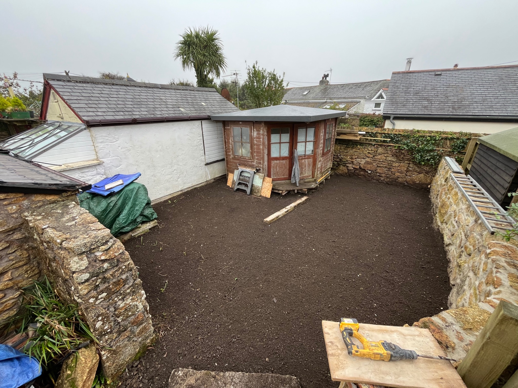 Przygotowany teren pod budowę altany ogrodowej. Ziemia wyrównana, widoczny sprzęt budowlany i elementy konstrukcyjne. Otoczenie stanowią domy i kamienne mury.