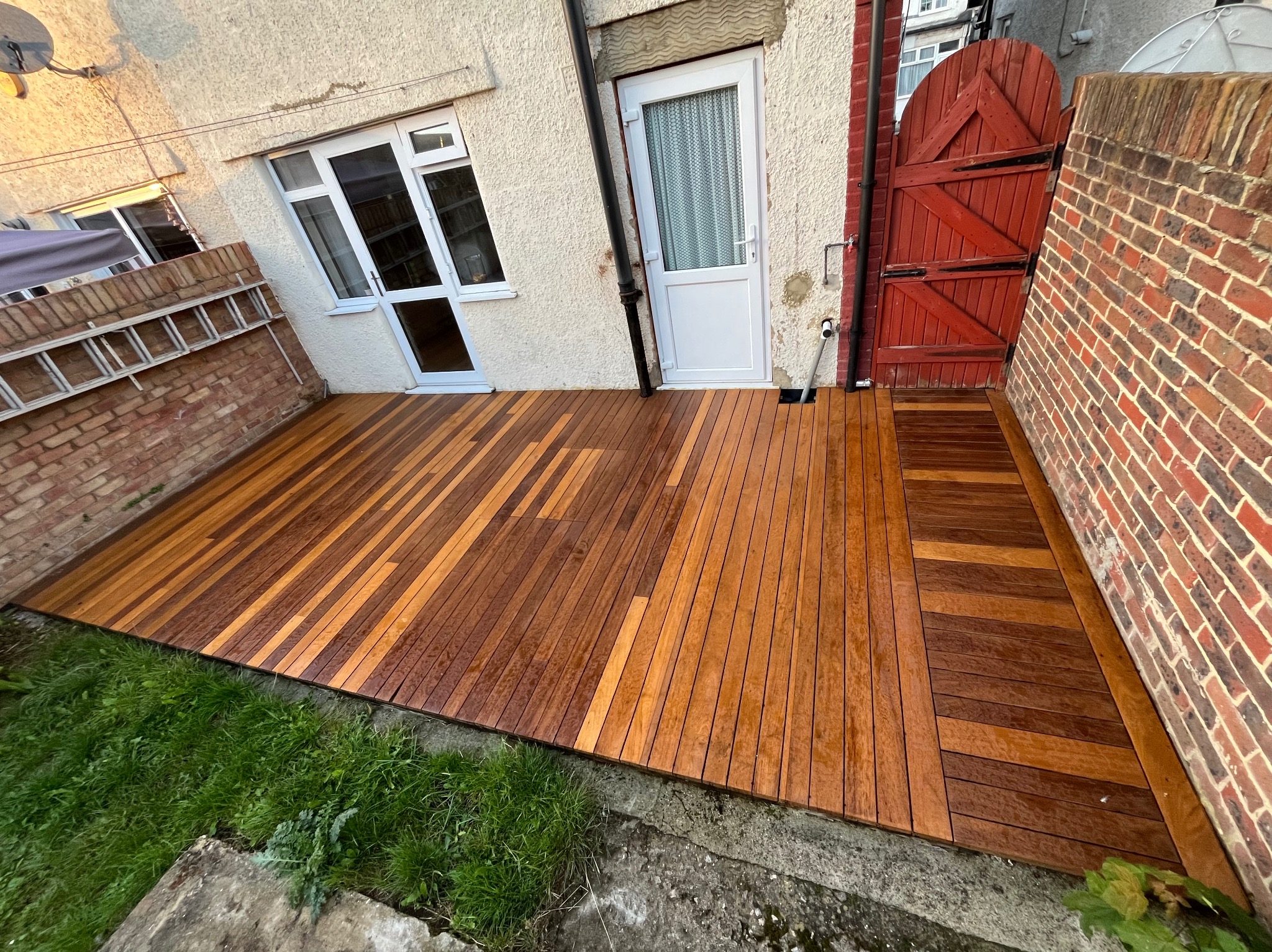 Nowy taras z desek kompozytowych w odcieniach brązu, przylegający do ściany budynku z oknem i drzwiami. Widoczna ceglana ściana i fragment trawnika.