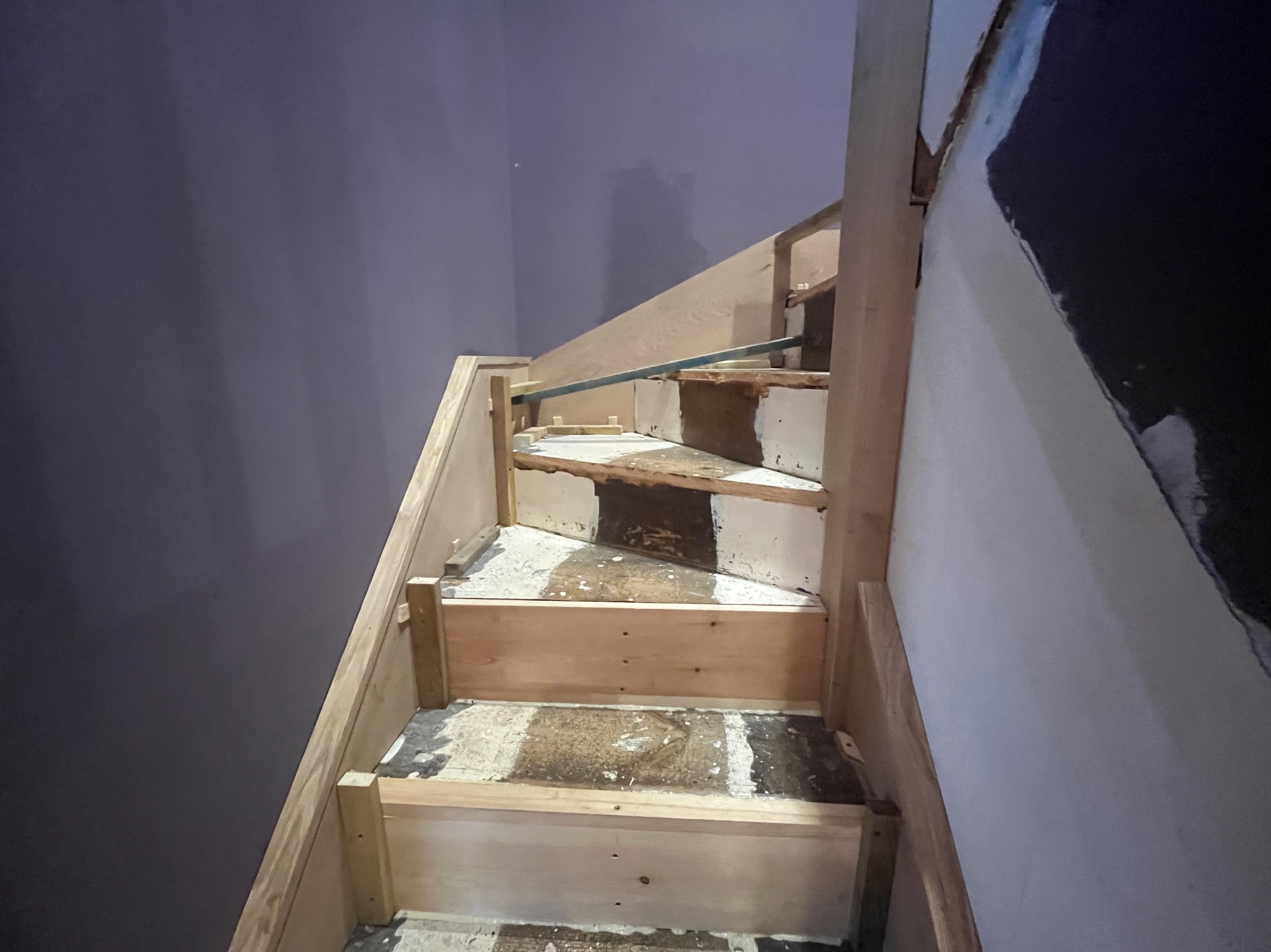 Drewniana konstrukcja schodów w trakcie tworzenia, widoczne elementy mocujące i surowy materiał. Ściana w tle pomalowana na ciemny kolor.