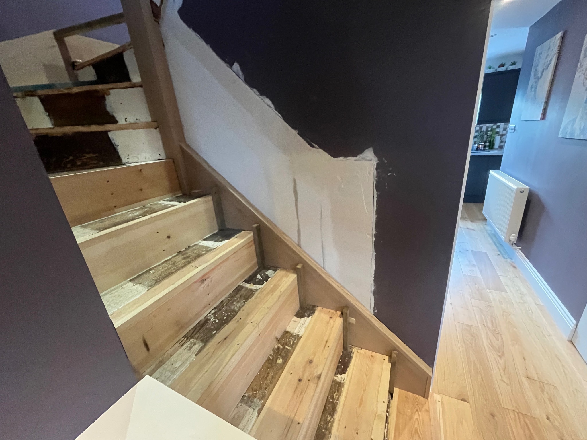 Drewniane schody w trakcie renowacji, z widocznymi surowymi stopniami i częściowo wykończoną ścianą w ciemnym kolorze. W tle widoczna kuchnia.