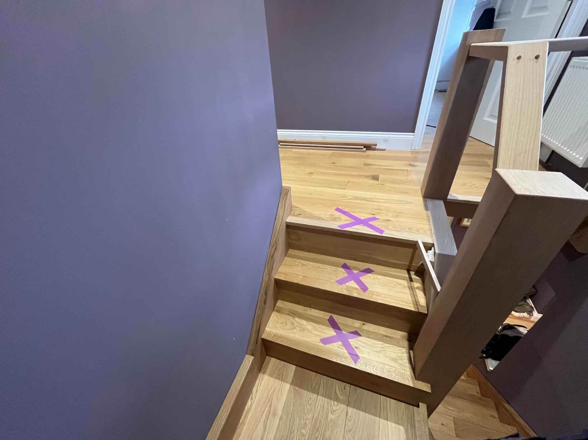 Drewniane schody z fioletowymi krzyżami, prowadzące do pokoju z podłogą z desek. Fragment konstrukcji schodów w jasnym drewnie. Ściana w kolorze fioletowym.