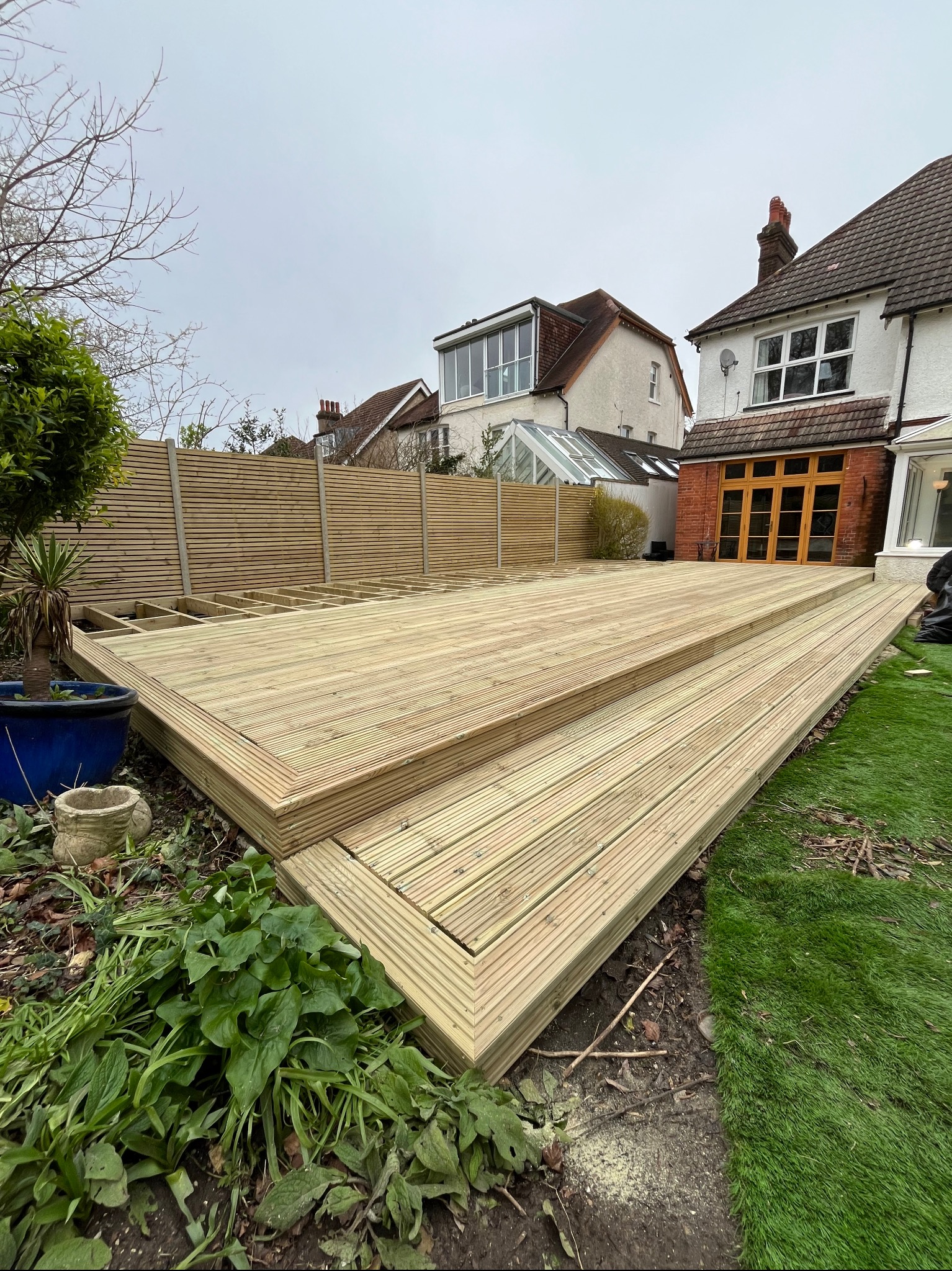 Nowy drewniany taras z dwoma stopniami, częściowo wykończony, z widoczną konstrukcją i ogrodzeniem w tle, na tle domu z ceglaną elewacją i ogrodem.