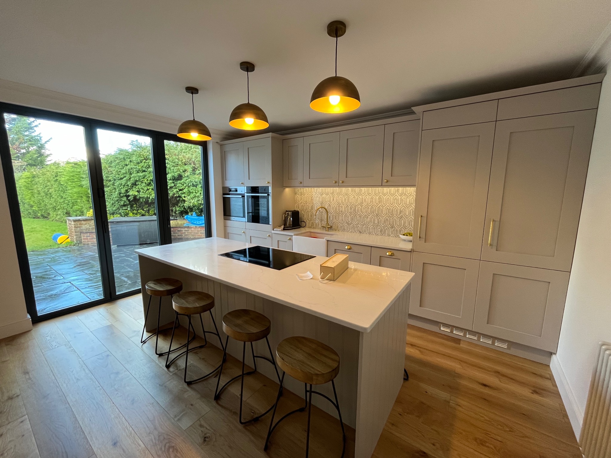 Elegancka, jasna kuchnia z wyspą i drewnianymi stołkami barowymi. Widoczne okno z widokiem na ogród i trzy złote lampy wiszące nad blatem. Nowoczesny design.
