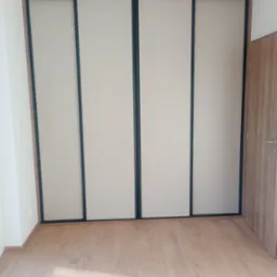 Czterodrzwiowa szafa wnękowa z przesuwnymi, beżowymi frontami i czarnymi obramowaniami, na tle drewnianej podłogi i fragmentu ściany z drzwiami.