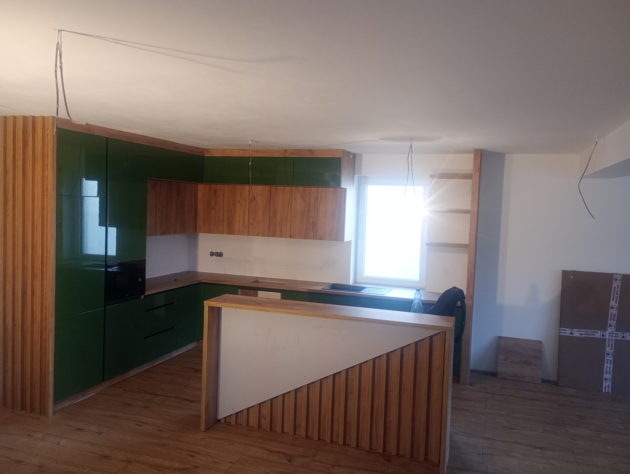 Nowoczesna kuchnia z zielonymi, lakierowanymi frontami szafek i drewnianym blatem. Widoczny barek z białym panelem i drewnianą okładziną. Częściowo wykończone wnętrze.