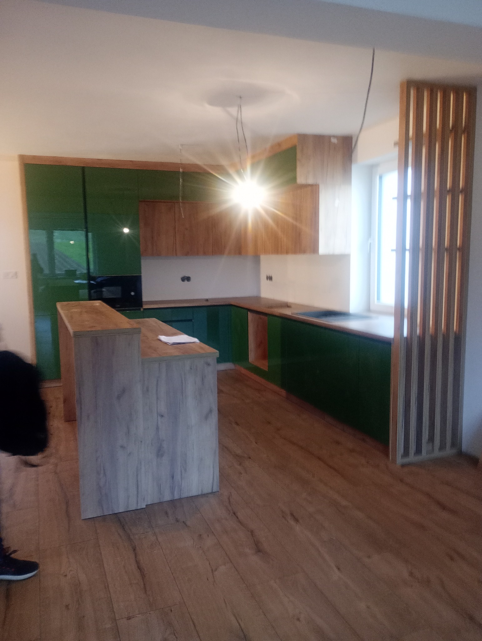 Nowoczesna kuchnia w odcieniach zieleni i drewna z wyspą, oświetlona jasnym światłem, z drewnianą ścianką działową i parkietem na podłodze.