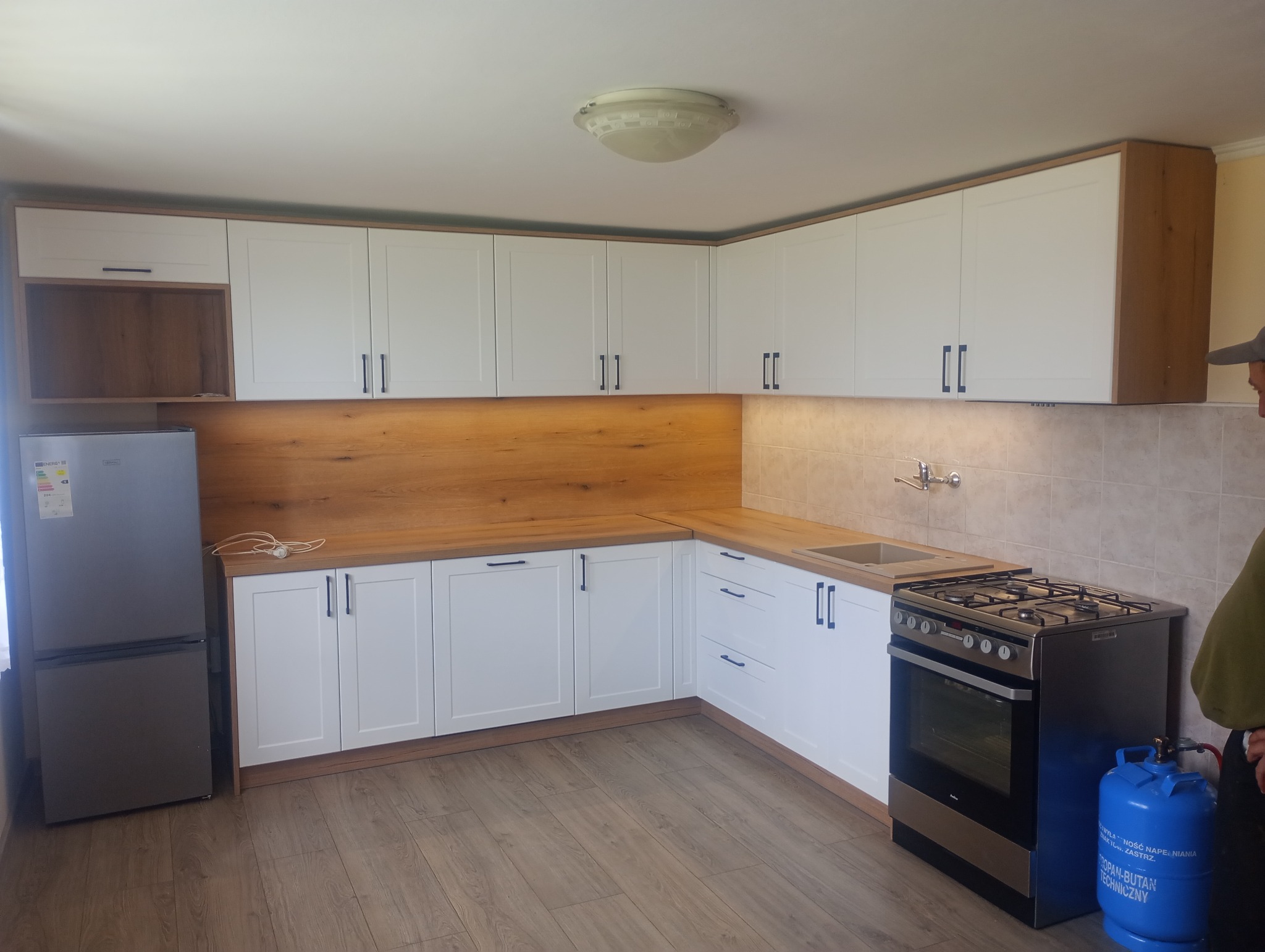 Jasna kuchnia w kształcie litery L z białymi frontami i drewnianym blatem. Na podłodze panele, widoczny fragment lodówki i kuchenki gazowej z butlą.