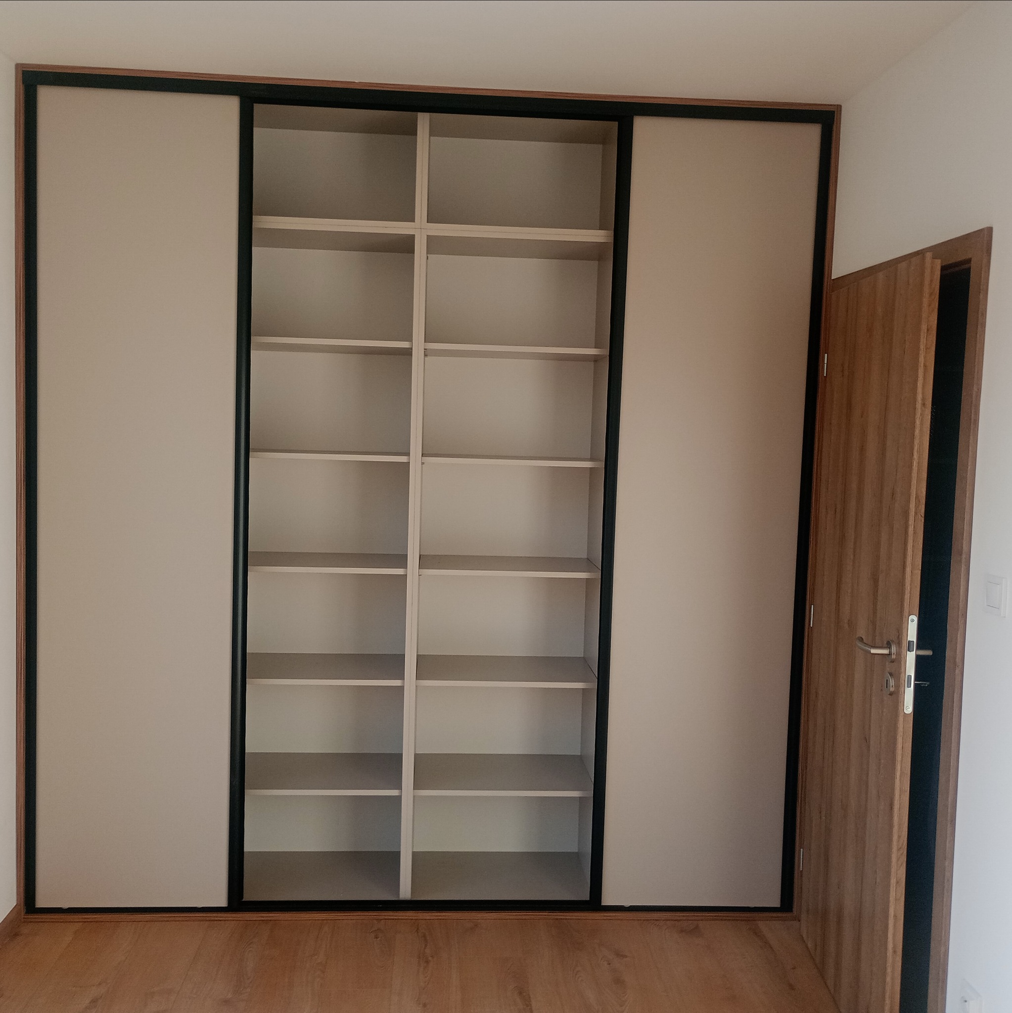 Jasna szafa wnękowa z przesuwnymi drzwiami i czarnymi obramowaniami, widok na puste półki, z boku drewniane drzwi wejściowe. Podłoga wyłożona panelami.