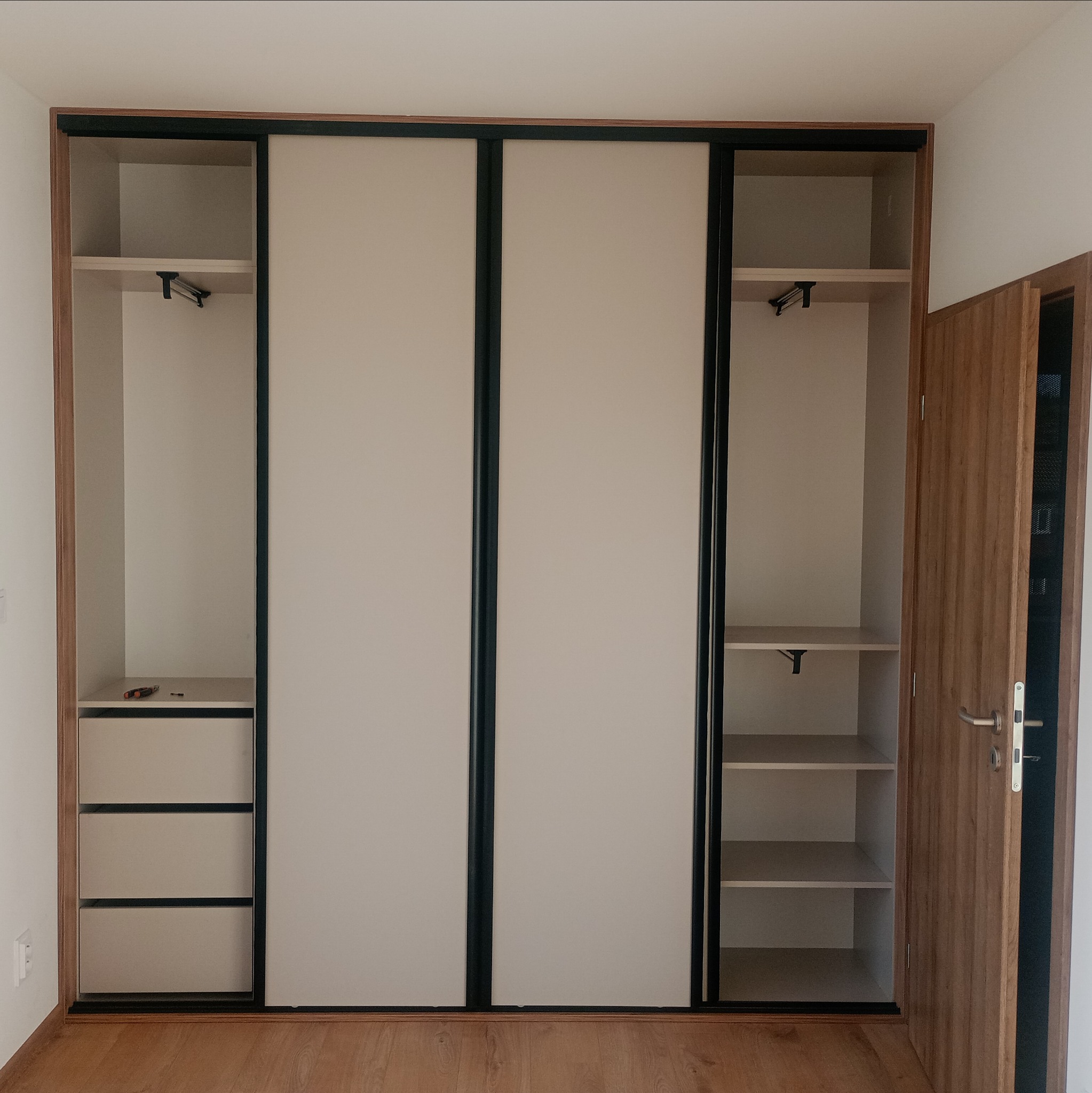 Jasna szafa wnękowa z przesuwnymi drzwiami i szufladami. Widoczne półki i drążki na ubrania. Otwarta drewniana szafa z drzwiami po prawej stronie.